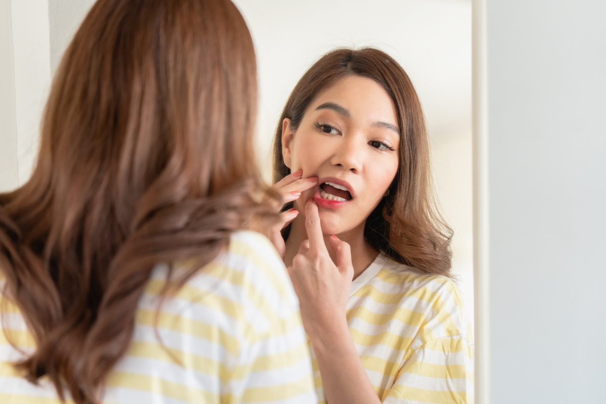Young woman looking into a mirror and pulling down her lower lip to check her gums or teeth. Concept of oral hygiene, dental health self-check, or mouth ulcer inspection.