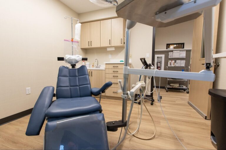 Dental treatment room with blue patient chair, overhead light, and cabinetry.