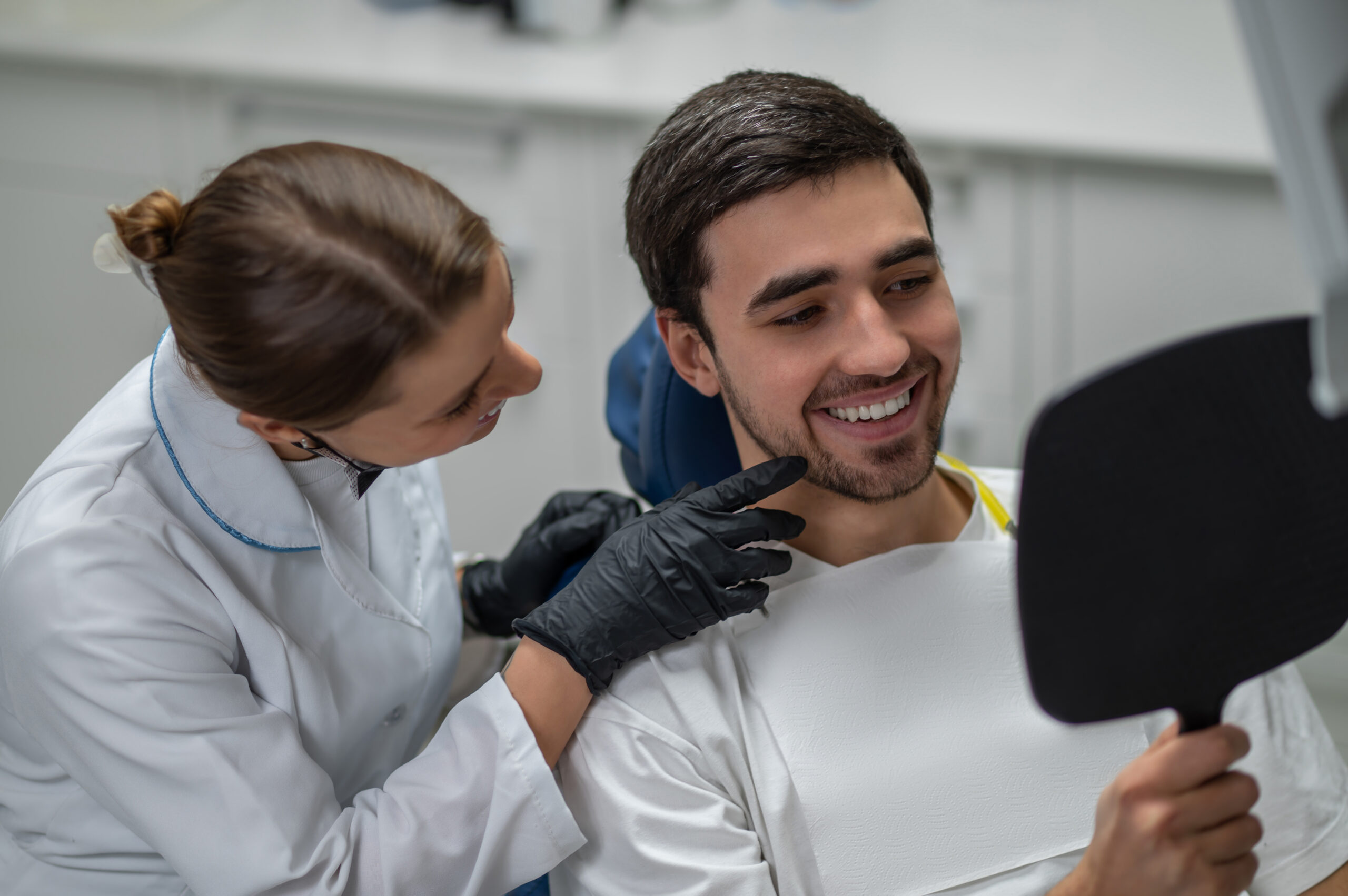 Smiling young man looking in hand mirror while female dentist pointing at his healthy white teeth after successful treatment in clinic