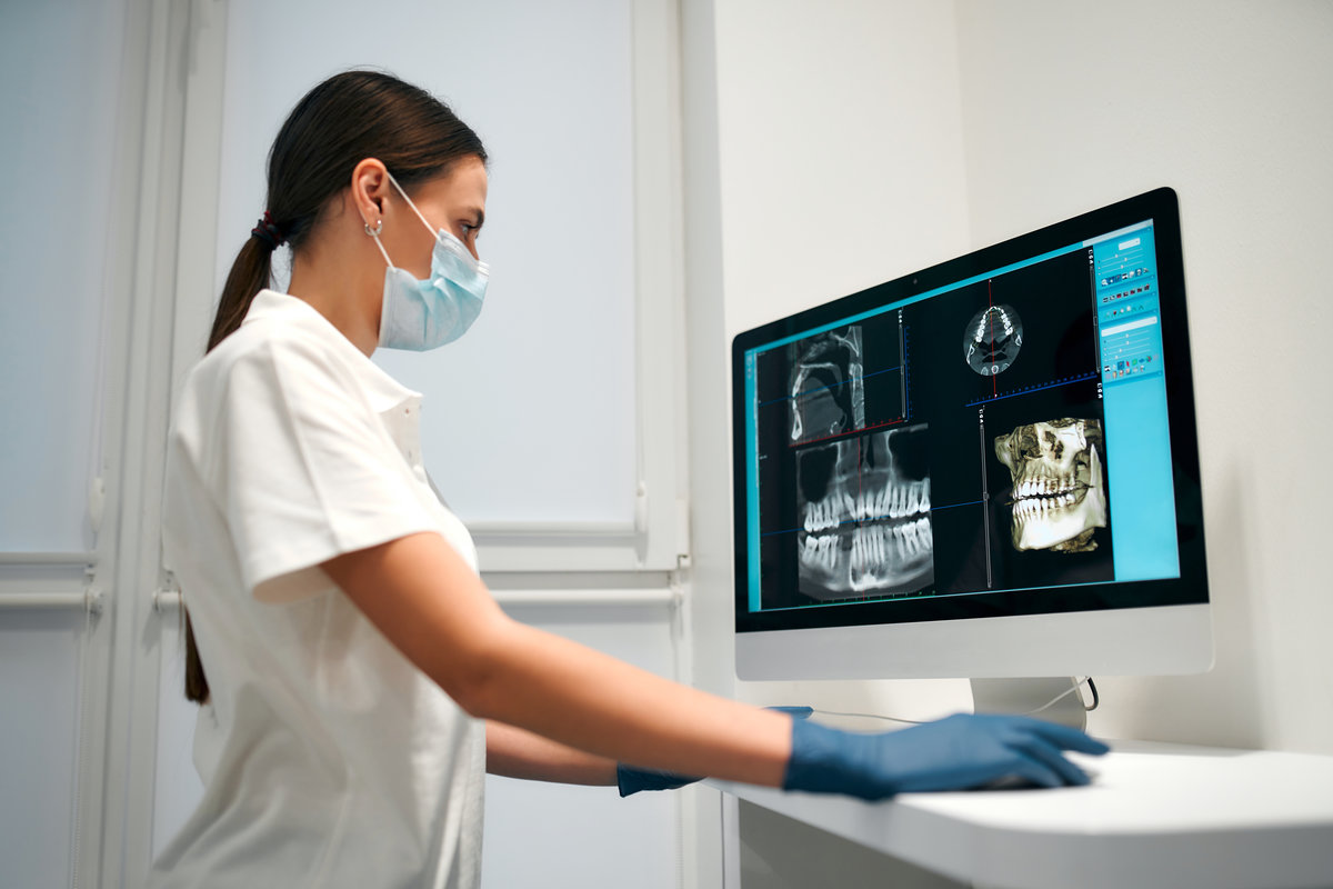 A young woman dentist in a medical mask examines an x-ray image on a computer in a dental office with modern equipment. Caries treatment. Dentistry and dental care.